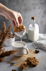 Hand dipping a homemade oatmeal cookie into a mug of milk. Milk drips from the cookie. Rustic breakfast setting with natural light and warm textures perfect for food and lifestyle themes. 