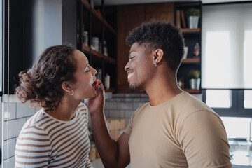 Naklejka premium A happy couple, Black and Latinx, shares a sweet moment in the kitchen. The man playfully feeds the woman a tomato, both smiling and enjoying their time together.