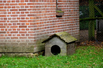 A view of a small hut, shack, shelter or barn made out of red brick and having an angled, tiled roof located next to a vast lawn, a wooden fence and a small wooden dog house seen in Poland