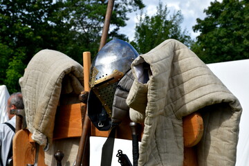 A close up on a set of medieval armor, including helmet, pike, jacket, and other weapons displayed next to some white cloth tent during a medieval fair organized in summer in Poland