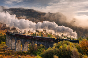 The morning train steams across the Glenfinnan Viaduct, its plume rising against the Highland mist. Golden light touches the hills, creating a scene of timeless beauty in Scotland’s heart.