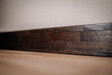 Close-up of a dark burnt wood baseboard against a textured white wall and wooden floor,...