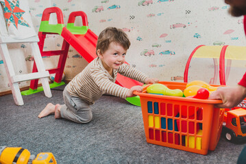 Naklejka premium Little boy 2 years old playing in the nursery at home. Shaking plastic toys out of the basket. Mess from a small child. Fun educational games. Independent play