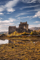 Eilean Donan Castle stands majestically amid autumn hues, its reflection shimmering in calm waters. Misty Highlands and golden trees create a scene of timeless beauty.
