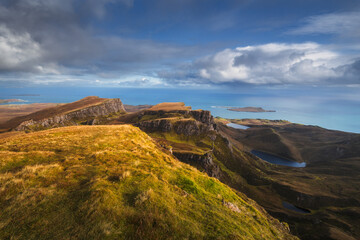 Autumn light bathes the Quiraing on Skye, casting long shadows over its dramatic cliffs and rolling hills. The landscape glows with golden hues, a breathtaking Highland scene.