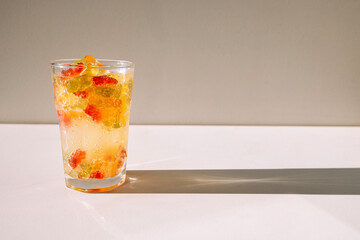 Close-up of a glass of mineral water with delicious marmalade bears and ice cubes on a white table. A cup of cool, bright, delicious sweet drink on a light background.