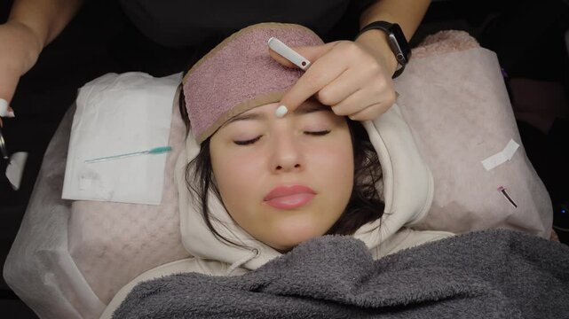 A beautician prepares a client for an eyelash extension session by adjusting under-eye patches while the client rests with eyes closed in a cozy salon setting.
