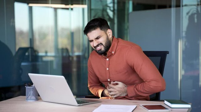 Upset businessman feels stomach pain while working on a laptop while sitting at workplace in business office. Sick worker has heartburn, gastritis or poisoning. Male suffers from spasms or constipated