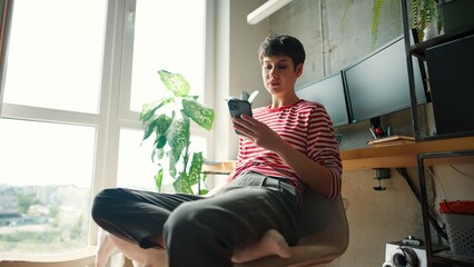 Charming Caucasian female with short dark hair enjoying day off. Sitting at desk in apartment. Scrolling social media with smartphone. Looking outside window while petting lovely dog.