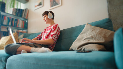 Obraz premium Bottom view of Caucasian woman remotely working or searching something on laptop. Female comfortably sitting on sofa in cosy living room. Wearing wireless headphones while pressing keys on laptop.