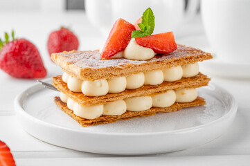 French dessert millefeuille with vanilla cream and fresh strawberries on a white plate on a white wooden background.
