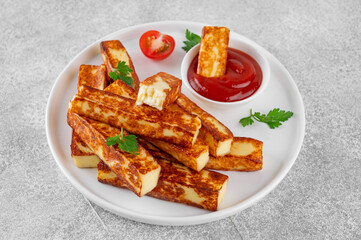 Fried halloumi cheese sticks with tomato sauce on a white plate on a concrete background.