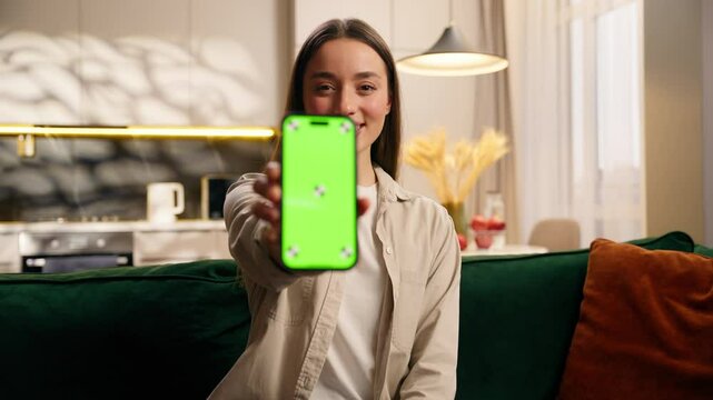Happy Caucasian woman girl female user showing smartphone mobile phone with green screen relaxing on sofa living room home color chroma key template layout for advertising smiling looking camera app