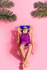 Photo of positive optimistic senior man wear bodysuit hold arms behind head relax rest chaise lounge isolated on pink color background