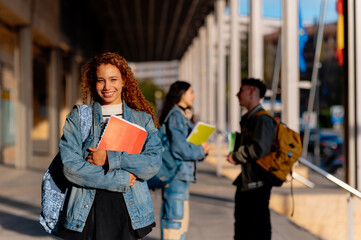 Portrait of a happy university student holding books on campus with classmates in the background,...