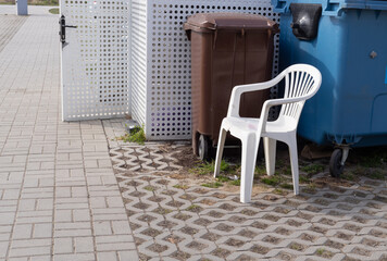 discarding a white plastic chair next to trash containers, outdoor scene showing a broken garden...