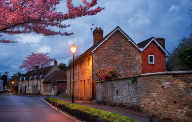 A charming evening in Iffley, Oxford, with historic Cotswold stone cottages, glowing streetlights, and vibrant cherry blossoms in spring season, UK