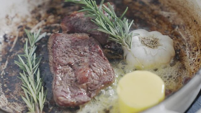 A piece of meat is being cooked in a pan with garlic and rosemary. The meat is browned and has a crispy texture. The garlic and rosemary add a savory flavor to the dish