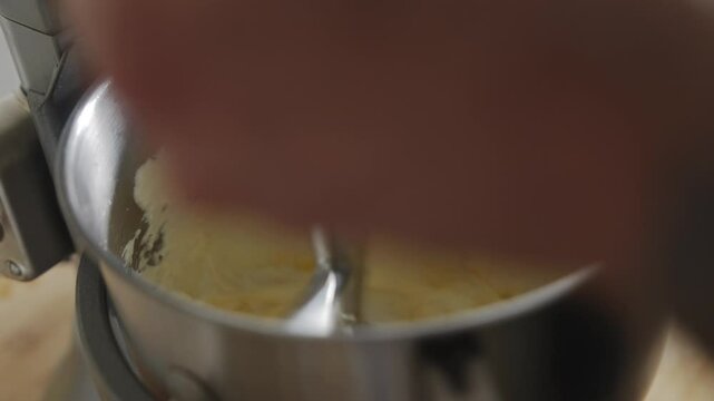 A person is mixing a batter in a bowl with a mixer. The batter is yellow and the mixer is silver