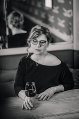Black and white photo of a woman with glasses sitting at a wooden table in a cafe, holding a glass of red wine and looking down pensively..