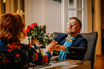 A man looks surprised as he receives a bouquet of roses from a woman during a Valentine’s dinner, suggesting a heartfelt yet slightly awkward gesture..