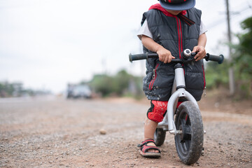 Naklejka premium Young Thai boy on a balance bike with muddy wheels and dirt-splattered clothes, focusing on the handlebars while sitting on a rough roadside path.