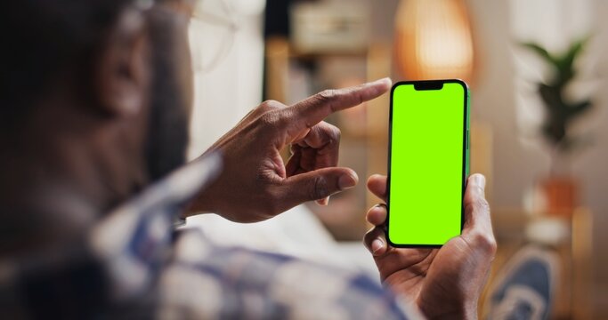 Back view of African American man holding phone with chroma key screen. Close up view of man looking at smartphone with green template display. Male using device with green mock up screen.
