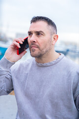 Caucasian man stands outdoors near water, talking on a smartphone with a serious look. His casual...