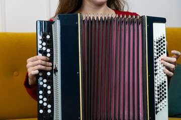 Woman musician playing a large black and white accordion with red bellows, sitting on a yellow sofa...