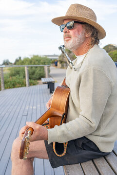 Aussie man playing a guitar and a harmonica sitting on a park bench