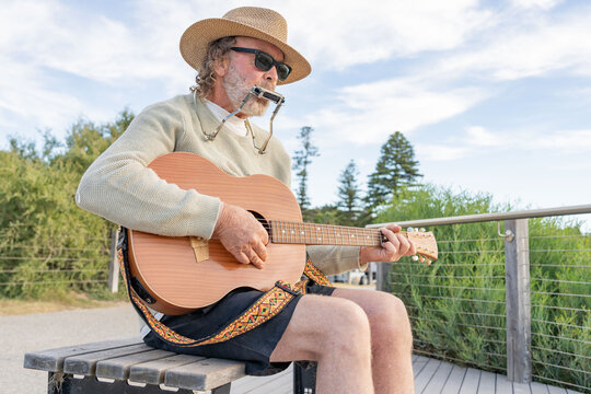 A busker wearing a hat playing a guitar and harmonica on a park bench