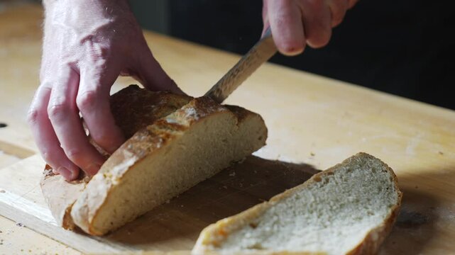 A person is cutting a piece of bread with a knife on a wooden cutting board. The bread is sliced in half, and the knife is positioned in the middle of the bread. Concept of simplicity and routine