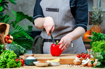 Chef shows how to cuts paprika. Cozy kitchen with wooden table, kitchenware, vegetables, herbs and ingredients for cooking. Healthy vegan eating, culinary, recipes, food blogging
