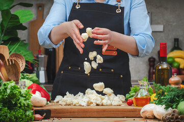 Chef pours fresh cauliflower florets, сozy kitchen with wooden table, kitchenware, vegetables, herbs and ingredients for cooking. Home food, cooking, recipes, blogging