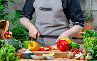 Chef shows how to cuts paprika. Cozy kitchen with wooden table, kitchenware, vegetables, herbs and ingredients for cooking. Healthy vegan eating, culinary, recipes, food blogging