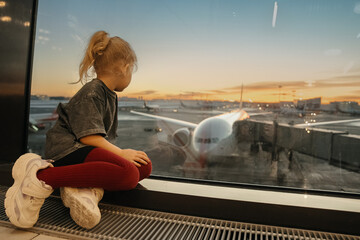 Child girl at the airport waiting for her flight looking out the window at the planes in the evening at sunset. Travel and children.