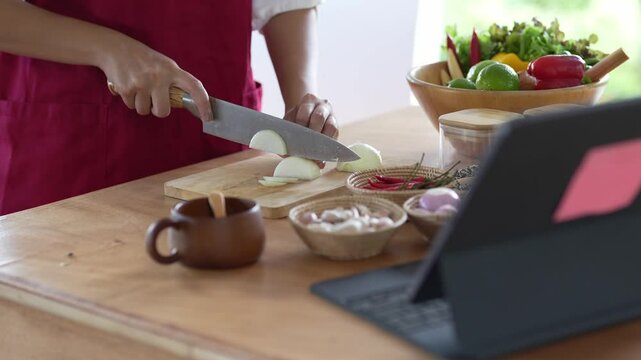A woman is cutting onions on a wooden cutting board in front of a laptop. The laptop is open and has a pink sticker on it. The woman is wearing a red apron