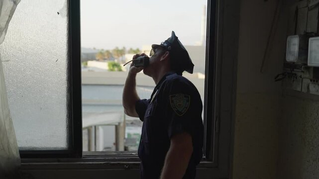 Police officer stands by window, looking out with a suspicious expression while reporting through his radio. Alertness, investigation, law enforcement, surveillance, security, public safety.
