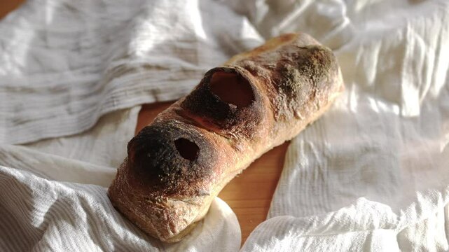 A piece of bread with holes in it is on a white cloth. The bread is burnt and has a dark crust. The cloth is white and has a striped pattern