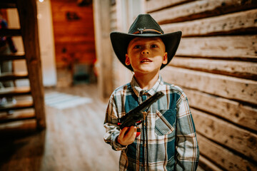 Smiling young boy in cowboy outfit with a black hat and plaid shirt holding a toy gun, standing indoors in a wooden rustic home environment..