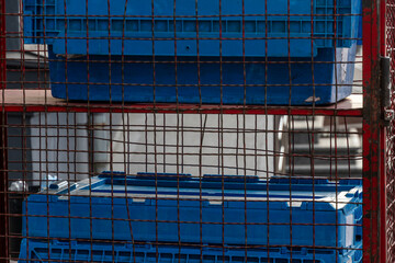 Stacked blue crates are placed behind a wire mesh barrier in a busy commercial area, with soft...