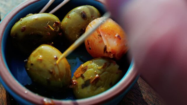 Close up of a hand picking up a green olive with a toothpick on a small bowl on a table