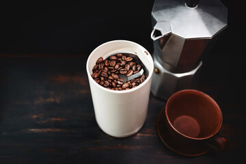 Roasted coffee beans in an electric coffee grinder on a wooden table with a moka pot and cup, coffee preparation