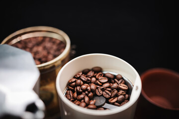 Roasted coffee beans in an electric coffee grinder on a table with a moka pot and cup, coffee preparation