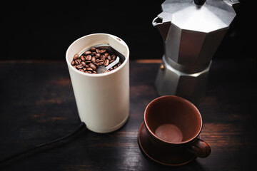 Roasted coffee beans in an electric coffee grinder on a wooden table with a moka pot and cup,...