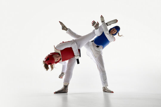 Two young women in blue and red taekwondo gear in dynamic motion, training against white background. Concept of martial arts, combat sport, strength, competition, power, endurance