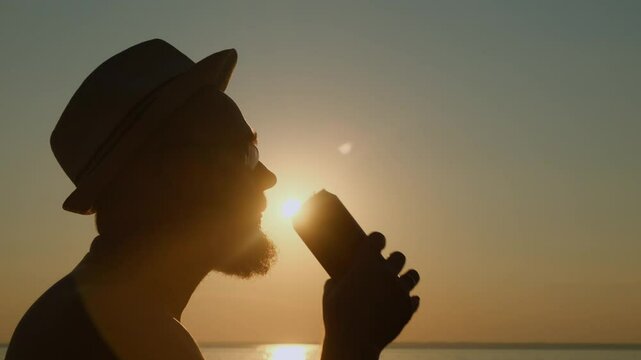 Adult bearded man in a hat drinking beer from a can by the sea at sunset. A refreshing sip of cold drink on a hot summer evening on vacation.