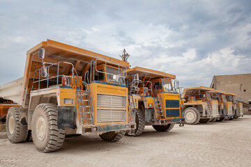 A row of large, heavy-duty mining dump trucks covered in dust, parked at a quarry under a cloudy sky. Industrial scene representing construction, mining, and heavy machinery operations. © dechevm