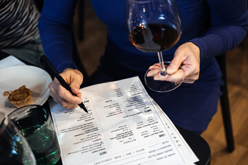Close-up of a woman filling out a tasting sheet while holding a glass of red wine during a...