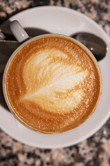 Top view of a creamy cappuccino with elegant latte art in a white ceramic cup with a spoon on a saucer, on a granite table. Coffee culture, creativity, barista skills concept and cozy cafe atmosphere